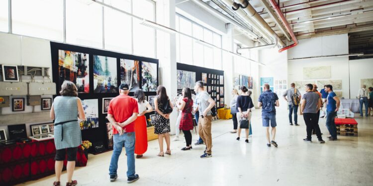 Geniş bir sanat galerisinde sergilenen tablolara bakan kalabalık sanatseverler.