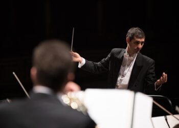 Siyah ceketli orkestra şefi bagetini tutarak orkestrayı yönetiyor.