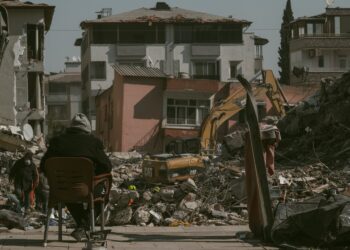 Yıkılmış binaların ve enkazın önünde oturan bir kişi ve iş makineleri.