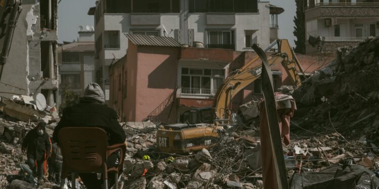 Yıkılmış binaların ve enkazın önünde oturan bir kişi ve iş makineleri.