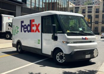 Şehir caddesinde seyreden yan tarafında elektrik sembolü bulunan FedEx kargo minibüsü.