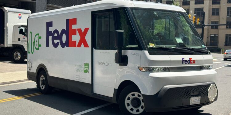 Şehir caddesinde seyreden yan tarafında elektrik sembolü bulunan FedEx kargo minibüsü.