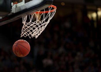 Kırmızı basketbol topu potaya doğru yaklaşırken salondaki kalabalık arka planda görünüyor.