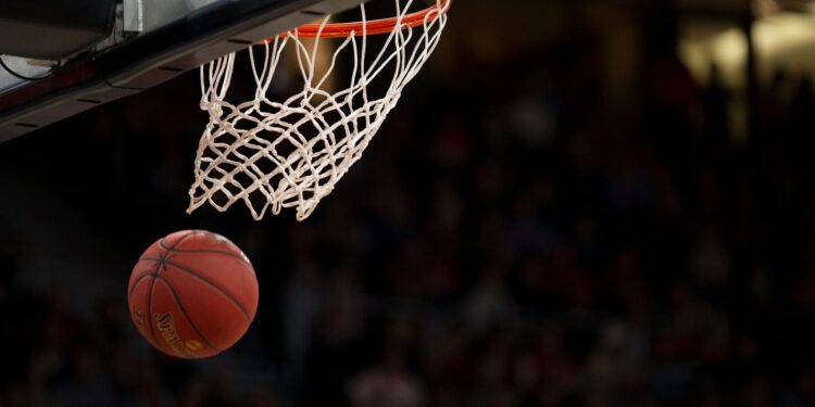 Kırmızı basketbol topu potaya doğru yaklaşırken salondaki kalabalık arka planda görünüyor.