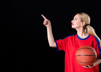 Sarı saçlı kırmızı formalı kadın elinde basketbol topuyla yukarıyı işaret ediyor.