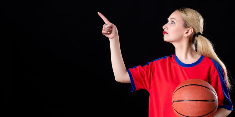 Sarı saçlı kırmızı formalı kadın elinde basketbol topuyla yukarıyı işaret ediyor.