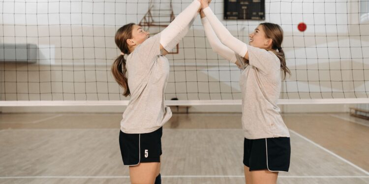 Kapalı salonda voleybol filesi önünde ellerini havada birleştiren iki genç kız.