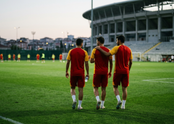 Stadyumda yürüyen üç futbolcu arkadan görülüyor, yeşil saha ve tribünler arka planda.