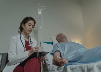 Yatakta yatan hastanın başında duran beyaz önlüklü kadın doktor not alıyor.