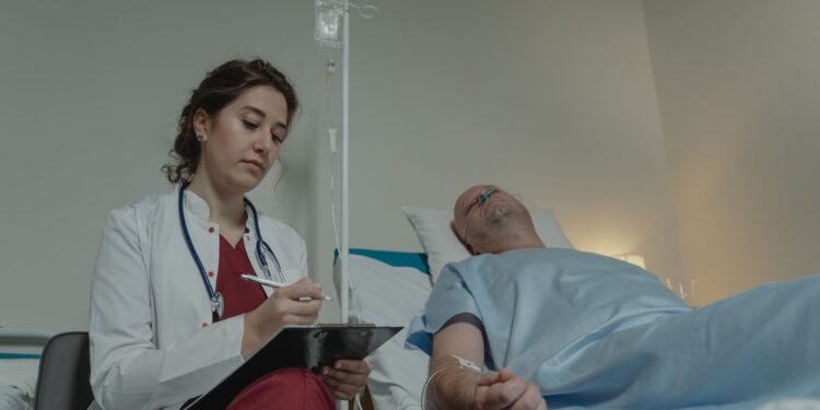 Yatakta yatan hastanın başında duran beyaz önlüklü kadın doktor not alıyor.