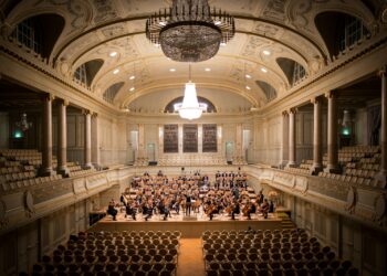 Dev avizelerle aydınlatılmış tarihi büyük konser salonunun sahnesinde orkestra performans sergiliyor.