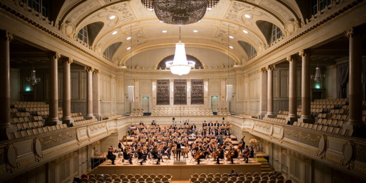 Dev avizelerle aydınlatılmış tarihi büyük konser salonunun sahnesinde orkestra performans sergiliyor.