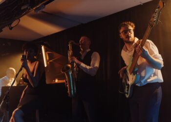 Loş sahnede şarkı söyleyen vokalist, saksafoncu ve bas gitarist.
