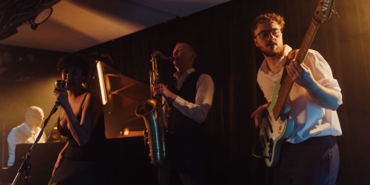 Loş sahnede şarkı söyleyen vokalist, saksafoncu ve bas gitarist.