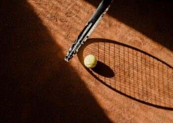 Toprak tenis kortunda bir raket, top ve gölgesi yer alıyor.