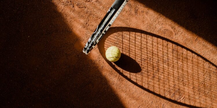 Toprak tenis kortunda bir raket, top ve gölgesi yer alıyor.